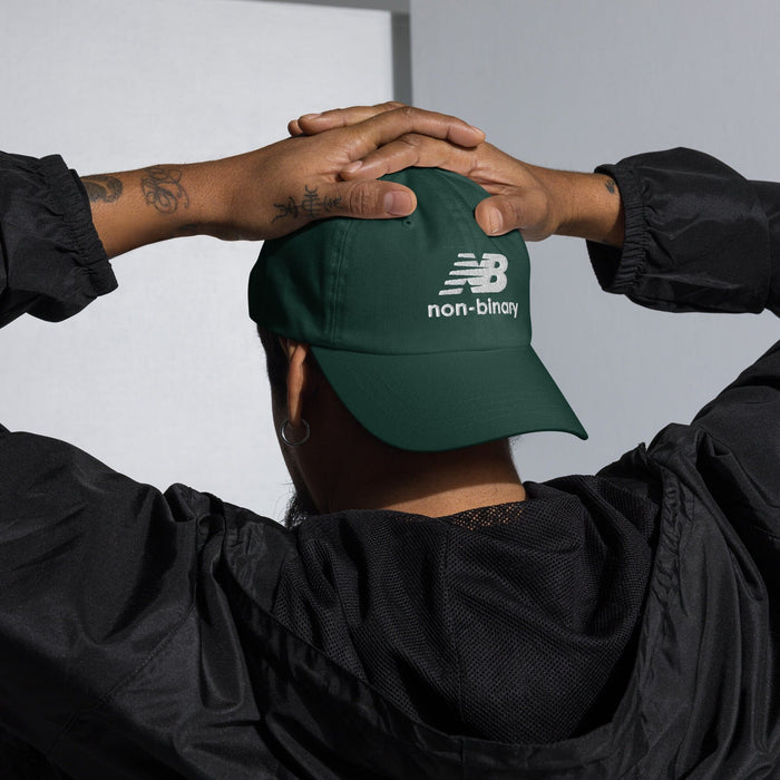 Person wearing a green cap with 'non-binary' text, hands clasped behind their head.