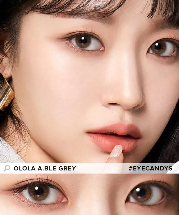 Close-up of a woman with smooth skin, subtle makeup, and Olola Able Grey color contact lens for dark eyes. She holds a finger to her lips and wears a gold earring. Image text: "Olola Able Grey #EYECANDYS".