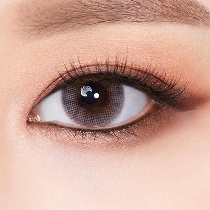 A close-up of an eye wearing Olola Amuse Grey by Olola, featuring long eyelashes, smooth skin, and soft brown eyeshadow. The iris is a striking grey shade, contrasting beautifully with natural makeup details.