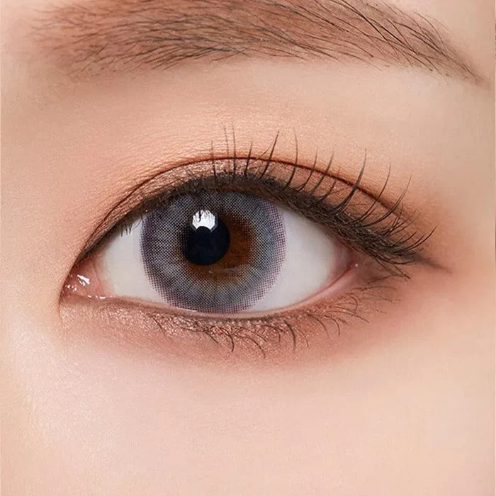 A close-up of a human eye with brown brows and long lashes wearing Olola Blow Midnight Grey by Olola, which gives dark eyes a striking blue-grey iris effect. The skin around the eye appears naturally smooth.