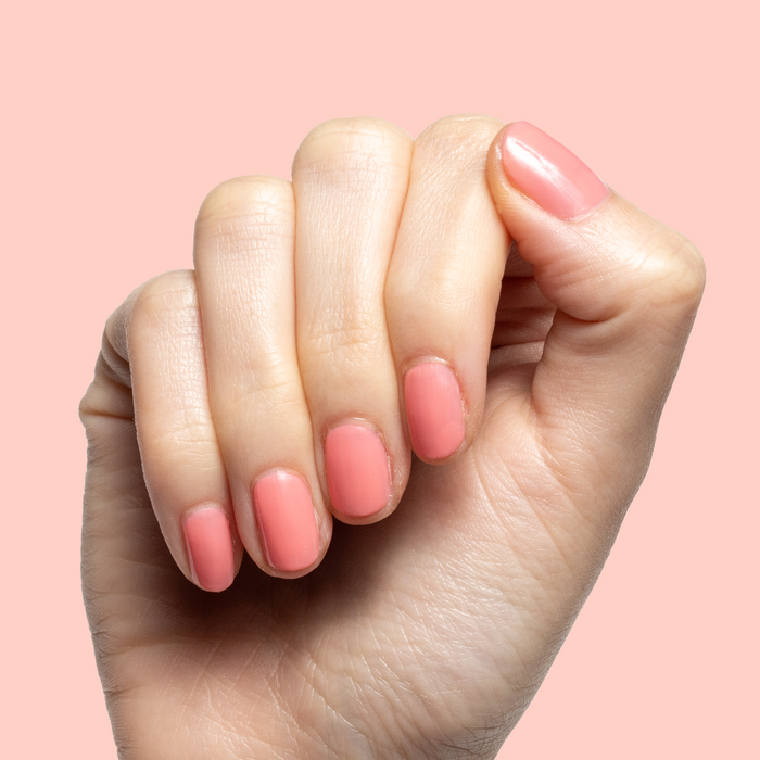 Hand with short natural nails painted in sheer coral pink using Papaya Gloss polish on soft pink background