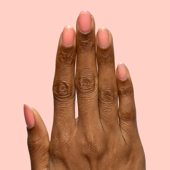Hand with medium natural nails painted in sheer coral pink using Papaya Gloss polish on soft peach background