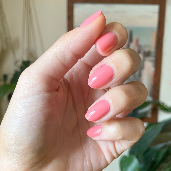 Hand with medium almond-shaped nails painted in glossy Papaya Gloss sheer coral pink polish indoors with blurred background