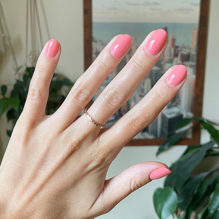 Hand with medium-length nails painted in vibrant coral pink using Papaya Gloss polish indoors with blurred city view background