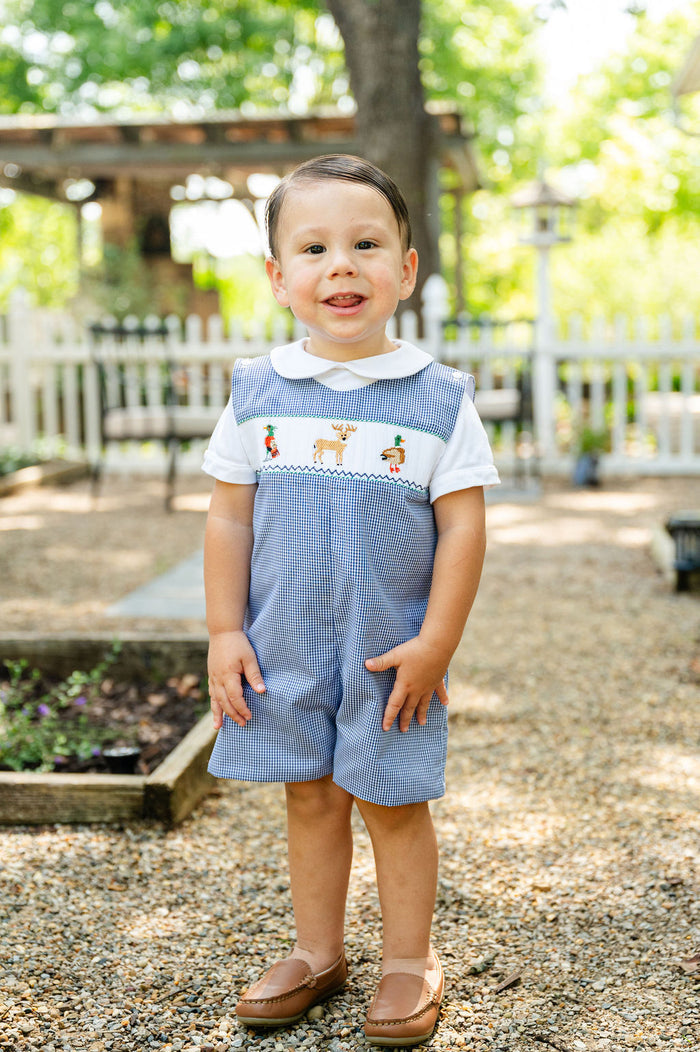 Baby boy standing outdoors wearing Smocked Hunting Jon Jon by Ruth and Ralph, blue gingham with smocked embroidery