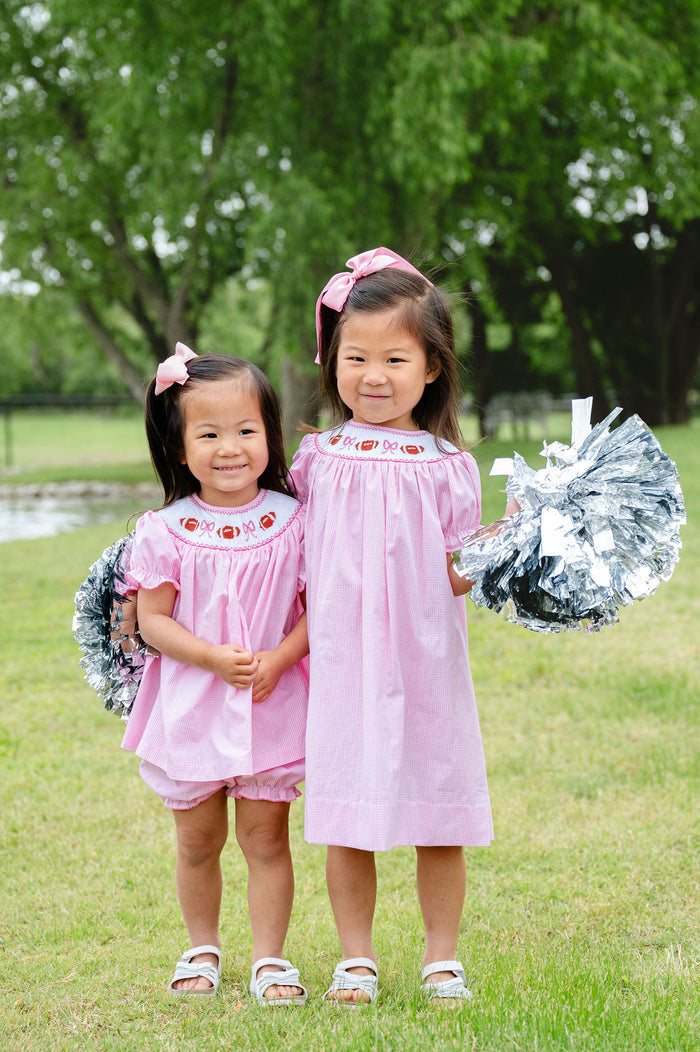 Two girls wearing matching pink gingham smocked football bloomer sets with hand-smocked footballs and bows