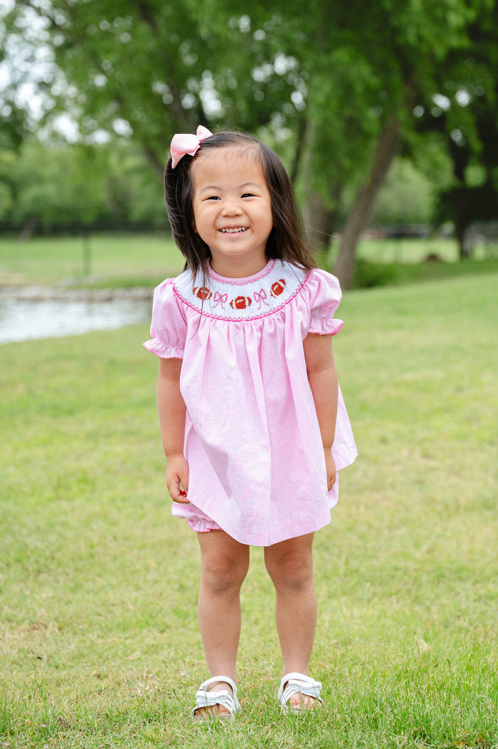 Toddler girl wearing a pink gingham smocked football bloomer set with a hand-smocked neckline of footballs and bows, standing outdoors