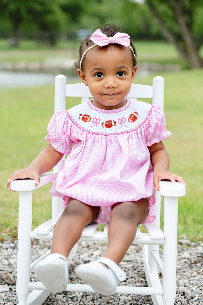 Baby girl wearing a pink gingham smocked football bubble with a hand-smocked neckline of footballs and bows, sitting in a white rocking chair