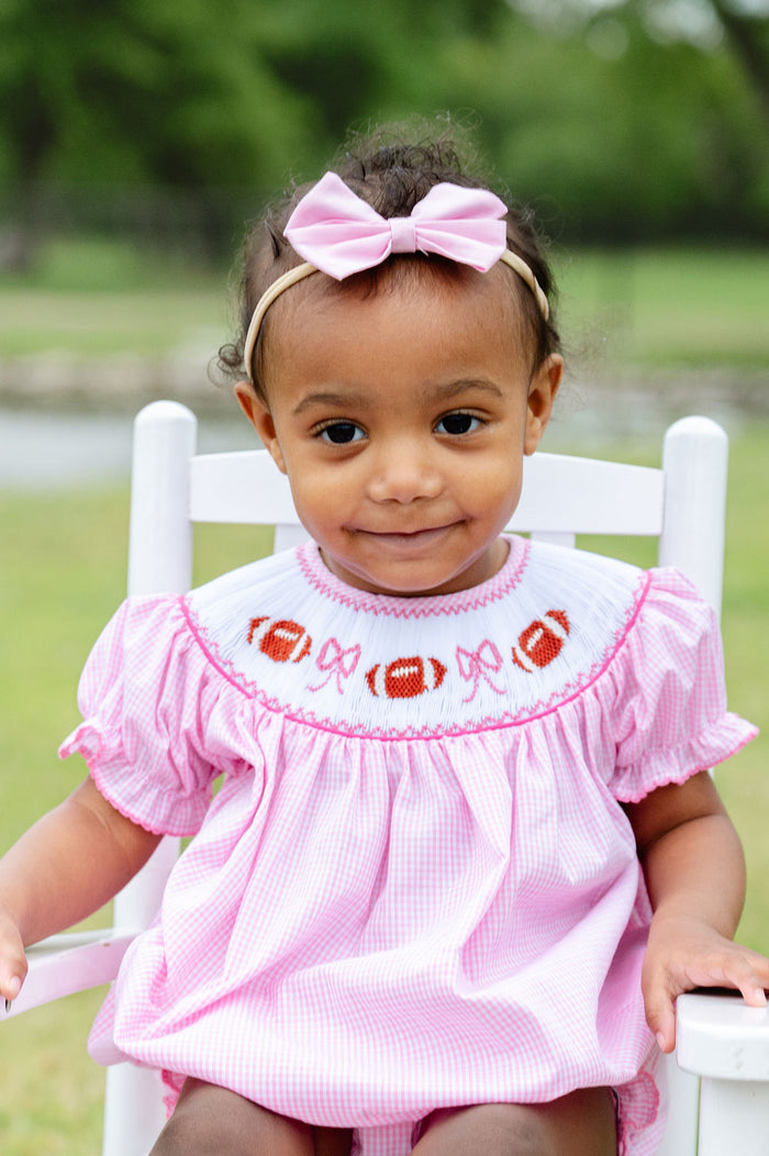 Smiling toddler girl in a pink gingham bubble with a hand-smocked football and bow neckline, seated outdoors