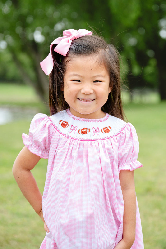 Toddler girl wearing a pink gingham smocked football bloomer set with a hand-smocked neckline of footballs and bows, standing outdoors