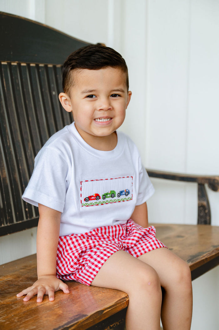 Boy sitting on wooden bench wearing Smocked Race Car T-Shirt and Shorts Set, red gingham shorts