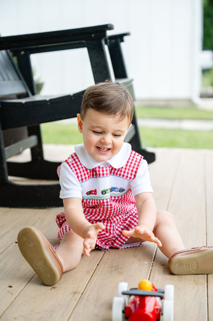 Baby boy sitting on porch playing with toy wagon, wearing Smocked Race Car Jon Jon by Ruth and Ralph, red gingham