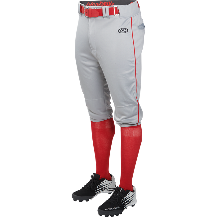 A person wears Rawlings Youth Launch Knicker Baseball Pants with red piping, a red belt, red high socks, and black cleats. Only the lower body is visible against a white background.