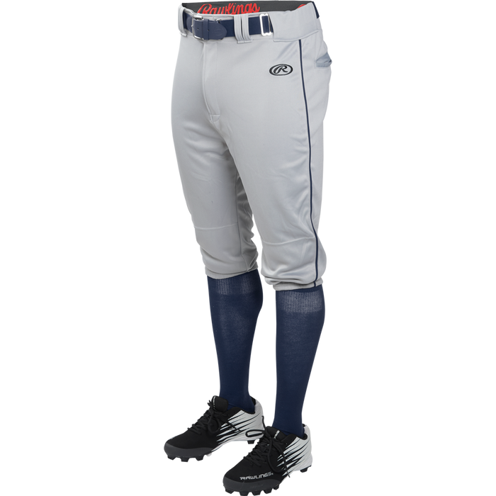A person wearing Rawlings Youth Launch Knicker Baseball Pants with Piping (YLNCHKPP), blue socks, and black cleats stands against a plain white background.
