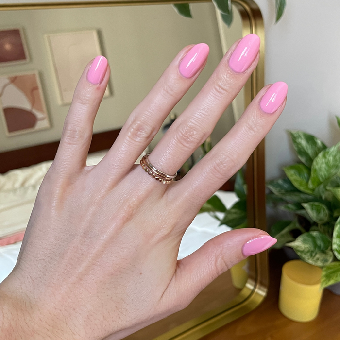 Hand displaying glossy nails painted in bright pink Saltwater Taffy gel polish with rings and home decor background