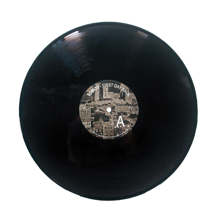 A black vinyl record by Somos with a center label showing a sepia cityscape, the text First Day Back, a large letter A, and a subtle Black Backpack motif on the right side.
