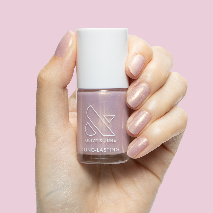Hand holding Star Baker nail polish bottle with sheer iridescent lavender gel polish on natural nails against pink background
