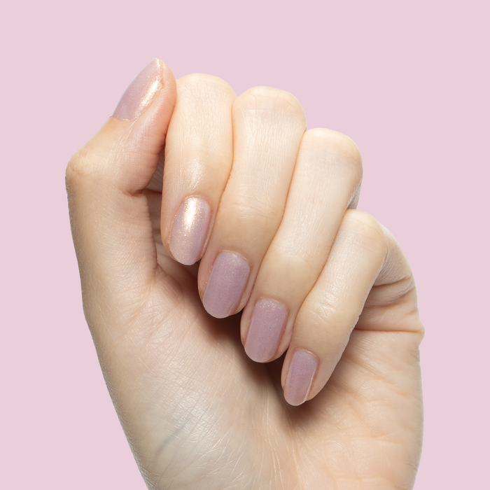 Hand with short nails polished in Star Baker sheer iridescent lavender gel polish against pale pink background