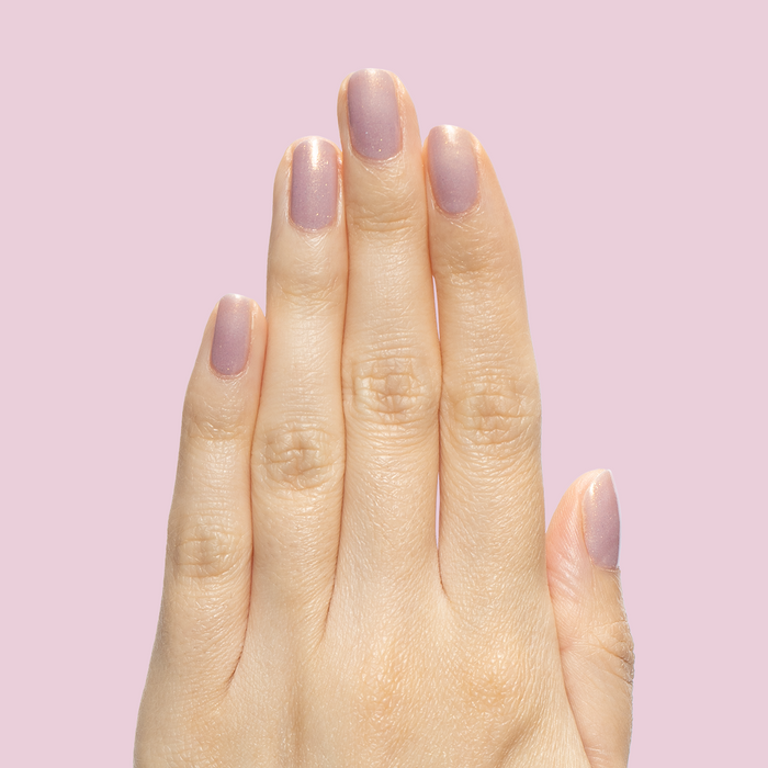 Hand with short rounded nails polished in Star Baker sheer iridescent lavender gel polish on pale pink background