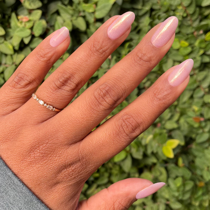 Hand with almond-shaped nails polished in Star Baker sheer iridescent lavender gel polish against natural green leaves background