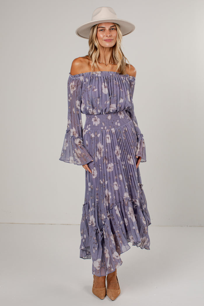 A woman modeling an off-the-shoulder midi dress in a dusty purple floral print, paired with a wide-brimmed cream hat and tan heels, against a plain light gray studio background.