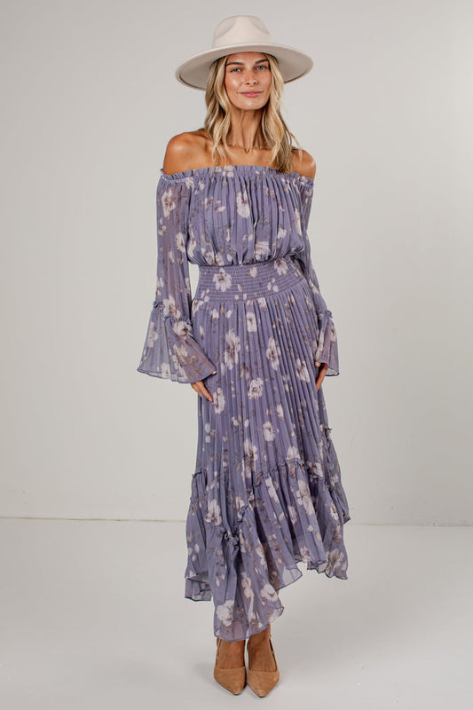 A woman modeling an off-the-shoulder midi dress in a dusty purple floral print, paired with a wide-brimmed cream hat and tan heels, against a plain light gray studio background.