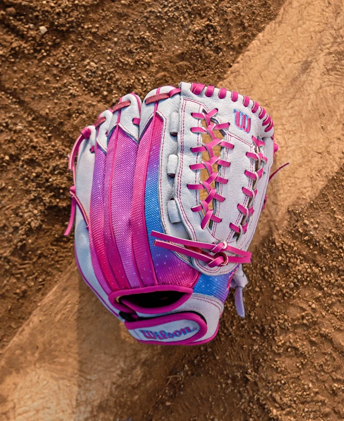 A Wilson Flash 12" Fastpitch Glove (WBW10398712) by Wilson in pink, purple, and light blue lies on a dirt infield with a white chalk baseline visible.